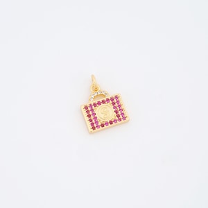 May include: A gold-colored charm in the shape of a handbag, encrusted with small, pink gemstones. The center features a gold-colored circle with the letter "S". The top handle is adorned with clear gemstones.