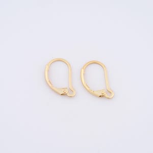 May include: Two gold-colored earring hooks are displayed against a white background. Each hook has a curved, elongated shape with a hinged clasp mechanism. The hooks are designed for securing earrings.