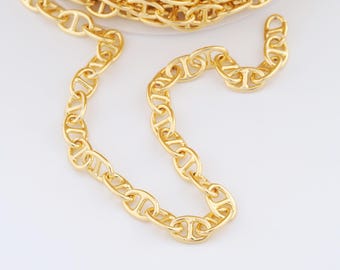 May include: Close-up of a gold-colored chain with a nautical anchor link design. The chain is coiled and spread out on a white surface, showcasing its shiny, metallic finish. The links are uniform and create a continuous pattern.