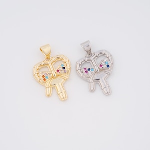 May include: Two pendants, one gold-toned and one silver-toned, each shaped like a stylized key with a heart-shaped bow. The bows are embellished with colorful gemstones. The pendants are displayed against a white background.