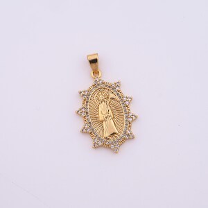 May include: Gold-colored pendant featuring a religious figure within an oval frame, surrounded by a starburst design. The pendant is adorned with small, clear gemstones and includes a loop for a chain. The background is plain white.