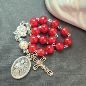 May include: A rosary with vibrant red beads and silver-toned accents. The rosary features a Padre Kolbe medal and a detailed crucifix. A silver rose and a pearl-like bead are also present.