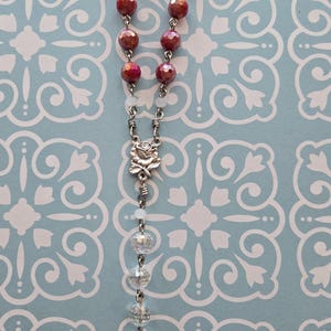 May include: A rosary featuring faceted deep red beads, clear crystal beads, and small white spacer beads connected by silver-toned metal links. A silver-toned metal rose centerpiece sits above a circular medal depicting a saint with the text Italy.