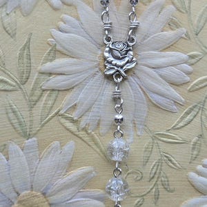 May include: A rosary with red and clear beads, connected by silver-toned metal links. The centerpiece features a rose design, and the pendant is an oval medal with a religious figure.