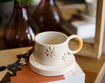 May include: A white ceramic teacup and saucer set with daisy accents. The teacup has a yellow handle and sits on a matching saucer. The set rests on a book with the word "KINFOLK" printed on the cover. Decorative vases and flowers are in the background.