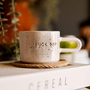 Può includere: Una tazza di ceramica macchiata con le parole "FUCK OFF" in rilievo sul davanti. La tazza ha un manico unico e poggia su un sottobicchiere intrecciato. La tazza è sopra un libro con la parola "CEREAL" stampata sul lato.