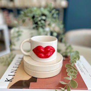 以下が含まれることがあります： 赤い唇のデザインが施された白いセラミックマグカップが、お揃いのソーサーのスタックの上に置かれています。マグカップにはカーブしたハンドルが付いています。アイテムは、表紙に「KINFOLK」という文字が印刷された本の上に置かれています。