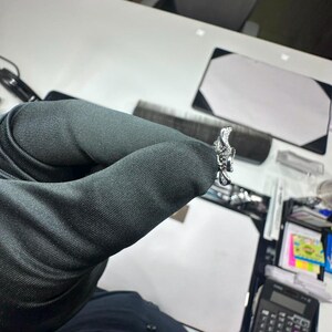 May include: A silver-toned ring featuring a detailed, textured floral or organic design with small, clear, sparkling stones embedded along the surface. The ring is held in a dark-colored gloved hand, showcasing its intricate metalwork and reflective finish.