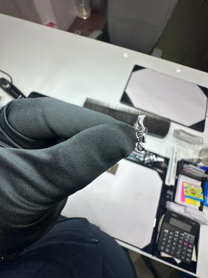 May include: A silver-toned ring featuring a detailed, textured floral or organic design with small, clear, sparkling stones embedded along the surface. The ring is held in a dark-colored gloved hand, showcasing its intricate metalwork and reflective finish.