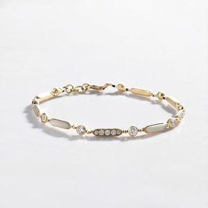 May include: A gold chain bracelet featuring alternating round and elongated links. The round links are set with clear stones, while the elongated links contain smooth white inlays. The bracelet includes a lobster claw clasp and a small extender chain.