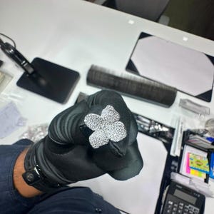 May include: A silver-toned ring featuring a large, four-petaled flower motif completely encrusted with small, clear, sparkling stones. The flower design is symmetrical, with each petal rounded and densely covered in reflective, brilliant-cut stones.