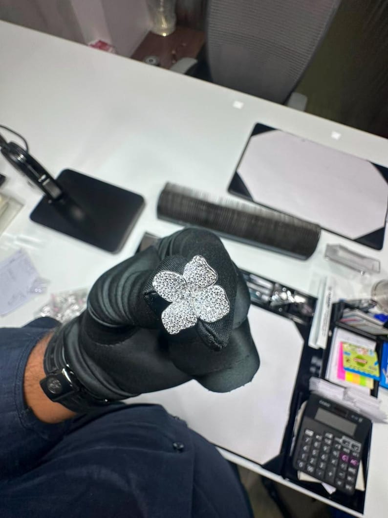 May include: A silver-toned ring featuring a large, four-petaled flower motif completely encrusted with small, clear, sparkling stones. The flower design is symmetrical, with each petal rounded and densely covered in reflective, brilliant-cut stones.