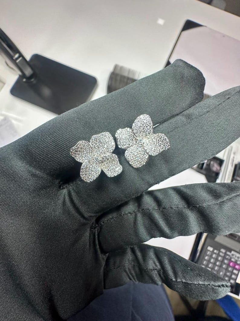 May include: A pair of silver-toned stud earrings, each shaped like a four-petaled flower. The surface of the petals is densely covered in small, clear, reflective stones that create a sparkling, textured appearance across the entire floral design.