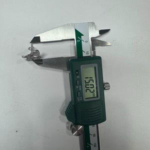 May include: A silver-toned butterfly-shaped earring with clear pav&eacute; stones being measured by a digital caliper. The display on the green and silver tool clearly shows a measurement of 15.02 mm, indicating the width of the jewelry piece.