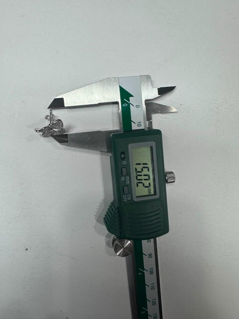 May include: A silver-toned butterfly-shaped earring with clear pav&eacute; stones being measured by a digital caliper. The display on the green and silver tool clearly shows a measurement of 15.02 mm, indicating the width of the jewelry piece.