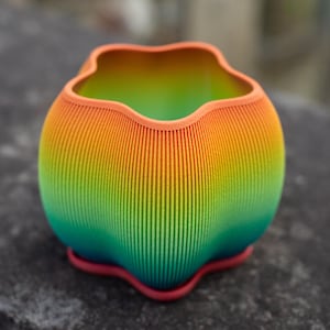 以下が含まれることがあります： ユニークな波型の縁を持つ、鮮やかな虹色のプランター。プランターはオレンジから黄色、緑、ターコイズへと変化します。テクスチャ加工されたリブ加工の表面を持ち、小さな赤いベースの上に置かれています。植物用の装飾品。