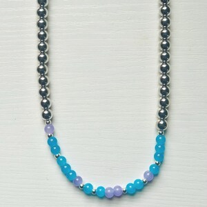 Op de afbeelding: Een ketting met zilverkleurige kralen aan elke kant, verbonden door lichtblauwe en lavendelkleurige kralen. De ketting wordt gepresenteerd tegen een witte achtergrond.