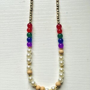 Peut inclure: Un collier à chaîne dorée avec des perles rouges, vertes, bleues et violettes formant un arc-en-ciel. Le collier alterne avec des fausses perles blanches, des perles en bois et des entretoises dorées.