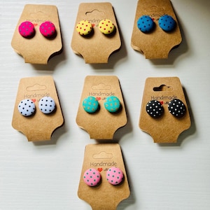 Puede incluir: Una colección de pendientes de botón hechos a mano en varios colores, cada uno con un estampado de lunares. Los pendientes se exhiben en tarjetas de cartón marrón. Las tarjetas tienen la palabra "Handmade" impresa.