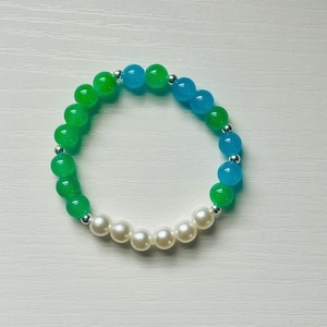 Può includere: Un braccialetto di perline con perline verdi, blu e bianche. Il braccialetto presenta una combinazione di perline rotonde e lucide nei toni del verde e del blu, con una sezione di perline di perle bianche. Distanziatori color argento separano le perline.