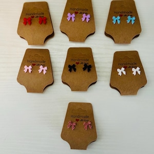 Pode incluir: Uma coleção de brincos de laço em várias cores, incluindo vermelho, roxo, azul, rosa, preto e branco. Cada par é exibido em um cartão marrom com o texto "Handmade with love".
