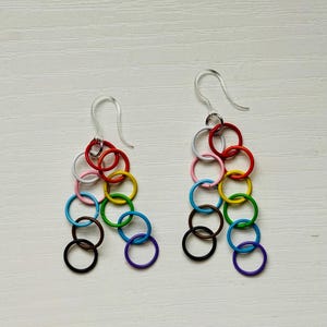 May include: A pair of earrings with a chainmail design, each composed of colourful interlocking rings. The earrings display a rainbow of colours, including red, orange, yellow, green, blue, purple, pink, white, brown, and black, suspended from silver-tone ear wires.