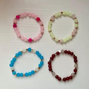 May include: Four beaded bracelets in shades of pink, green, blue, and red. Each bracelet features round beads and gold-colored spacers. The bracelets are arranged on a white surface, showcasing their colors and designs.