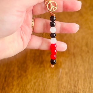 May include: A beaded keychain featuring a gold-colored peace sign charm and a series of black, white, and red beads. The keychain is attached to a gold-colored ring. The beads are approximately 0.5 inches in diameter.