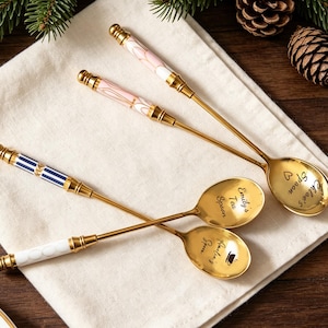 Puede incluir: Cuatro cucharas doradas con mangos decorativos y cuencos grabados. Los mangos presentan diseños rosas, blancos y azules. Los cuencos están grabados con nombres y las palabras "Tea Spoon". Las cucharas descansan sobre un paño blanco.