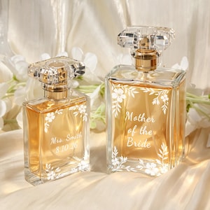 May include: Two rectangular glass perfume bottles with gold-toned liquid and clear crystal-like stoppers. The smaller bottle features the text Mrs. Smith 8.10.26, while the larger bottle displays the text Mother of the Bride, both with white floral engravings.