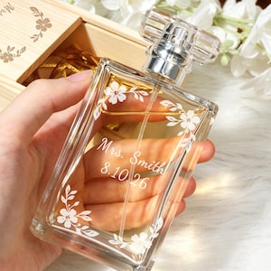 May include: A rectangular glass perfume bottle featuring a clear liquid, a silver spray nozzle, and a transparent cap. The front of the bottle is etched with white floral vine designs and the text Mrs. Smith 8.10.26 in a cursive script font.