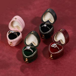 May include: An assortment of heart-shaped ring boxes in various colors, including pink, black, green, and burgundy. Each box is open, revealing a diamond ring inside. The boxes are arranged on a red surface.