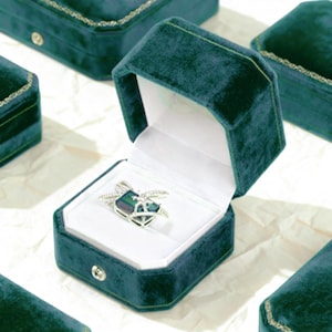 May include: A teal velvet ring box with a white interior holds a silver ring with a geometric design and a dark green and blue stone. Other teal boxes are in the background.