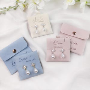 Könnte beinhalten: Ohrringe mit floralem Design und tropfenförmigen Perlen werden auf personalisierten Karten und in kleinen Beuteln präsentiert. Die Karten sind mit Namen wie "Amelie" und "Olivia" beschriftet, darunter das Wort "Brautjungfer". Die Beutel sind in Pastellfarben gehalten.