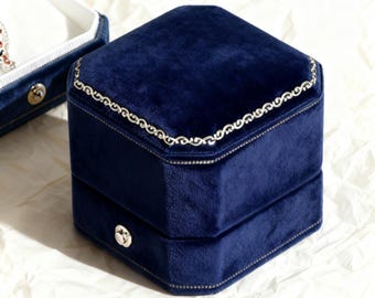 May include: A navy blue velvet jewelry box with a silver decorative trim around the lid. The box is hexagonal and has a silver clasp. Another open box is visible in the background.