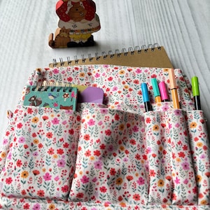 May include: A fabric organizer pouch featuring a white background with a dense floral pattern in shades of pink, red, orange, and green. The pouch has multiple exterior pockets containing a small spiral notebook, pens, and markers.