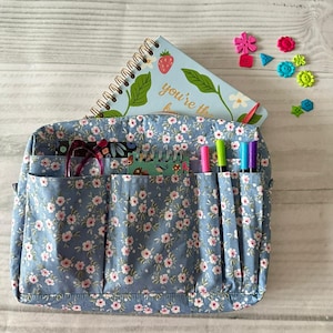 Può includere: Una borsa organizer blu con stampa floreale con più tasche, contenente un taccuino con la scritta "you're the" e penne colorate. Abbellimenti decorativi a forma di fiore sono sparsi intorno alla borsa.