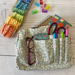May include: A floral print craft organizer bag with multiple pockets, holding scissors, pens, and a notebook. Beside it are colorful sewing clips and a selection of ruffled ribbons in shades of orange, yellow, green, blue, and purple. A pair of glasses rests in a pocket.