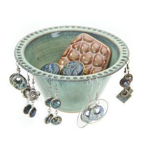 May include: A light green ceramic bowl with a decorative rim, used as a jewelry holder. The bowl is filled with various earrings and a small ceramic tile with a brown and white pattern.