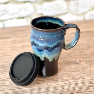 May include: A ceramic travel mug with a black lid. The mug features a blue and teal glaze with a brown base and handle. The lid is made of black silicone. The mug is designed for hot or cold beverages.