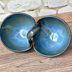 May include: Two connected ceramic bowls with a handle, featuring a glossy blue and teal glaze. The bowls have a circular shape and a dark rim. The handle connects the two bowls, creating a unique serving piece.