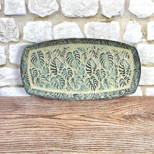 May include: A rectangular ceramic tray with rounded corners featuring a repeating embossed pattern of monstera leaves. The surface has a light cream-colored base glaze with dark green accents highlighting the textured leaf details throughout.