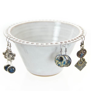 May include: White ceramic bowl with a scalloped edge and holes around the rim, used as an earring holder. Three pairs of earrings are displayed in the bowl, including a pair of silver flower earrings, a pair of blue and white patterned earrings, and a pair of brown and gold earrings.