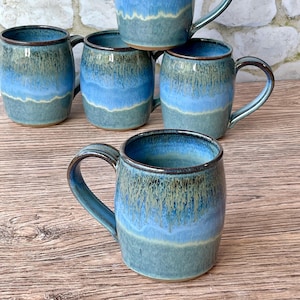 May include: A collection of handmade ceramic mugs with a unique glaze. The mugs feature a gradient of blue and green hues, with a dark brown rim and handle. The mugs are cylindrical with a curved handle, perfect for enjoying hot beverages.