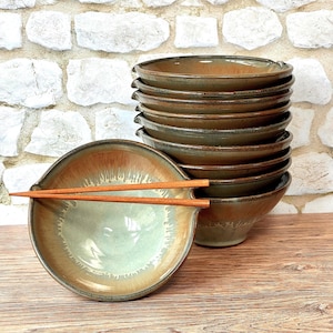 May include: A stack of ten brown and green ceramic bowls with a light green glaze. The bowls are stacked on a wooden surface. A single bowl with a light green glaze and brown accents is in the foreground with a pair of wooden chopsticks.