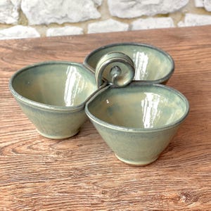 May include: A three-section ceramic serving dish featuring a glossy, light sage green glaze with subtle blue undertones. The bowls are connected by a central, spiraled ceramic handle, creating a unified piece with smooth, rounded edges.