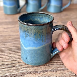 May include: A handmade ceramic mug with a blue and turquoise glaze. The mug has a dark brown rim and handle. Several similar mugs are visible in the background, suggesting a set. The mug is held by a person.