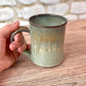 May include: A handmade ceramic mug with a green and brown glaze. The mug has a cylindrical shape with a handle and a dripped glaze effect. The mug is held by a hand, showcasing its size and design.