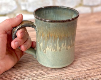 May include: A handmade ceramic mug with a green and brown glaze. The mug has a cylindrical shape with a handle and a dripped glaze effect. The mug is held by a hand, showcasing its size and design.