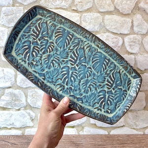 May include: A rectangular ceramic tray featuring a repeating embossed monstera leaf pattern. The surface is finished with a glossy glaze in shades of deep blue and light teal, with dark brown accents highlighting the textured leaf details.